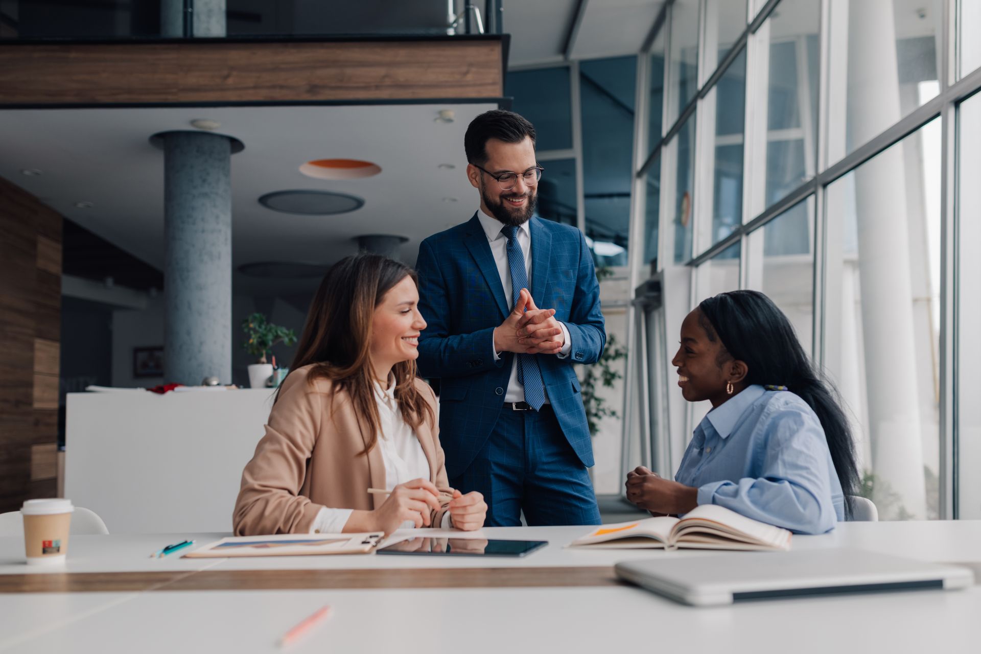 Smiling manager is standing and explaining business strategy to two female employees sitting at a desk in a modern office, creating a positive and collaborative work environment