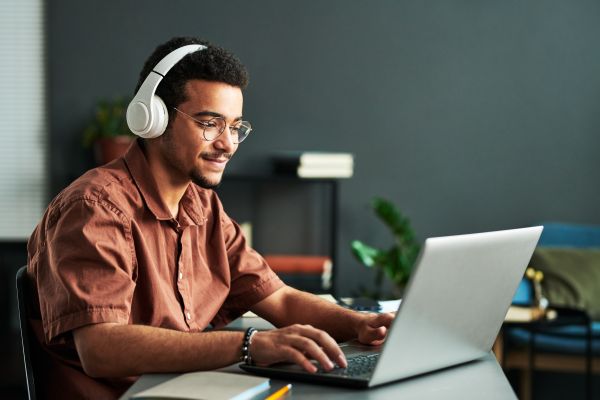 personalized_online_learning_adult Young smiling man in headphones typing on laptop keyboard while sitting by workplace and taking part in online webinar or lesson